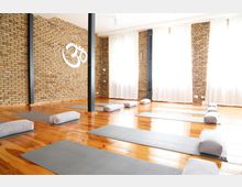 Heller Yoga-Raum mit Holzboden, grauen Yogamatten und Kissen. Die Ziegelwände sind mit einem großen weißen „Om“-Symbol dekoriert, und helle Vorhänge bedecken die Fenster.