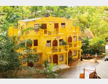 Gelbes, mehrstöckiges Gebäude mit Balkonen und Holztüren, umgeben von üppiger tropischer Vegetation. Im Vordergrund eine gepflasterte Fläche mit einigen Menschen, die sich in der Nähe eines Sitzbereichs aufhalten.