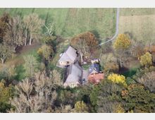 Luftaufnahme eines ländlichen Gehöfts mit mehreren Häusern mit roten Ziegeldächern und Reetdächern, umgeben von Wiesen, Bäumen und einem schmalen Weg. Die Umgebung besteht aus dichtem Grün und herbstlich gefärbten Bäumen.