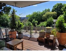 Eine Terrasse mit Holzboden und Geländer aus Metall, dekoriert mit Pflanzkübeln, Rankpflanzen und einem kleinen Tisch mit Sitzmöbeln. Im Hintergrund sind Bäume, Sträucher und ein Bahngleis mit einem Graffiti-bedeckten Zug zu sehen.