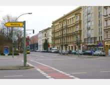 Straßenszene in einem Stadtviertel mit mehrstöckigen, historischen Gebäuden auf der rechten Seite, einer geparkten Autoreihe und einem gelben Ortsschild mit der Aufschrift „Wedding“. Im Vordergrund befinden sich eine Kreuzung mit Fußgängerüberweg und Radwegmarkierungen.