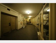 Ein schmaler, länglicher Raum mit schlichtem, funktionalem Design, gekennzeichnet durch beige Wände, einfache Türen und Glasvitrinen entlang der Wände. Der Raum ist mit mehreren Deckenlampen beleuchtet und erinnert an einen Bunker oder einen alten Krankenhaus- bzw. Lagereinrichtungsbereich.