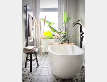 Modernes Badezimmer mit freistehender weißer Badewanne vor einem großen Fenster mit halbtransparenten Vorhängen. Pflanzen, ein dunkler Hocker mit gefalteten Handtüchern und florale Fliesen verleihen dem Raum eine natürliche und stilvolle Atmosphäre.