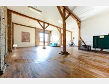 Großer, heller Raum mit Holzboden und sichtbaren Holzbalken sowie teilweise freiliegendem Ziegelmauerwerk. Im Raum befinden sich ein Sofa, eine Tafel, ein kleiner Tisch und eine Lampe, mit viel offener Fläche und großen Fenstern, die Tageslicht einlassen.