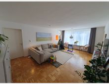 Helles Wohnzimmer mit Parkettboden, einer grauen Eckcouch, einem großen Fenster mit weißen Vorhängen und einem Esstisch mit zwei Rattansesseln. Im Raum befinden sich Kinder-Spielzeug und Zimmerpflanzen, ein Bild hängt an der Wand.