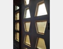 Wand mit geometrischen, sechseckigen Fensterausschnitten, die mit strukturiertem, lichtdurchlässigem Glas gefüllt sind und warmes Licht in den Raum lassen. Die Umgebung wirkt aus Beton und Stein gefertigt.