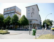 Straßenkreuzung in einer Stadt mit Geschäftsgebäuden im Hintergrund, darunter ein fünfstöckiges Wohn- und Geschäftshaus mit einem kunstvollen Wandmuster. Im Vordergrund befinden sich ein Fußweg, ein bepflanztes Beet und eine grüne Müllbox, umgeben von mehreren Bäumen.