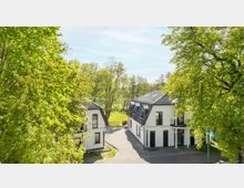 Zwei moderne, weiß gestrichene Gebäude mit schwarzen Dächern befinden sich in einer parkähnlichen Umgebung, umgeben von hohen, grünen Bäumen. Im Hintergrund ist eine weitläufige Wiese sichtbar.