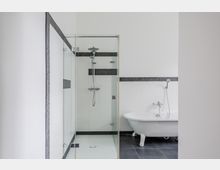 Modernes Badezimmer mit bodengleicher Glasdusche und großer freistehender Badewanne; weiße Wände werden durch dunkle Fliesen am Boden und eine schmale horizontale Fliesenbordüre akzentuiert.