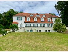 Weiße Villa mit rotem Ziegeldach und blauen Fensterläden, umgeben von einer gepflegten Rasenfläche und hohen Bäumen. Das Gebäude hat mehrere Etagen, halbrunde Dachfenster und eine leicht erhöhte Lage mit Treppen an der Seite.