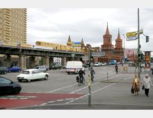 Straßenszene in Berlin mit der Oberbaumbrücke im Hintergrund, einer roten Backsteinbrücke mit Türmen, über die ein gelber U-Bahn-Zug fährt. Im Vordergrund befinden sich eine belebte Kreuzung mit Autos, Radfahrern und Fußgängern sowie Straßenschilder, darunter "Mühlenstraße".
