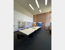 Konferenzraum mit einem rechteckigen weißen Tisch in der Mitte, umgeben von schwarzen Stühlen. Im Hintergrund befinden sich Infotafeln, Roll-Up-Banner und eine Flipchart vor einer Holzvertäfelung; die Deckenbeleuchtung besteht aus langen, rechteckigen Leuchten.
