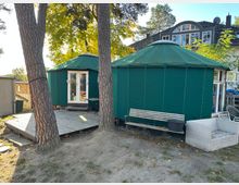 Zwei grüne, zeltartige Rundbauten mit Holzeingängen stehen auf einem sandigen Grundstück, umgeben von Bäumen. Im Vordergrund befinden sich ein Holzdeck, eine Parkbank und Gartenmöbel, im Hintergrund ist ein modernes Wohnhaus mit großen Fenstern sichtbar.