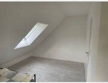 Minimalistisch eingerichtetes Dachzimmer mit weiß gestrichenen Wänden, einer Dachschräge mit einem schrägen Dachfenster und hellem Laminatboden. Am linken Bildrand ist ein Teil eines Betts mit gemustertem Bettbezug zu sehen.