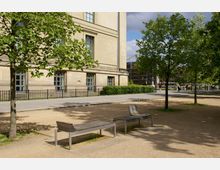 Eine städtische Parkanlage mit Schotterboden, schattenspendenden Bäumen und mehreren modernen Metallbänken. Im Hintergrund ein historisches Sandsteingebäude mit hohen Fenstern und ein Gehweg, hinter dem vereinzelt Personen vorbeigehen.