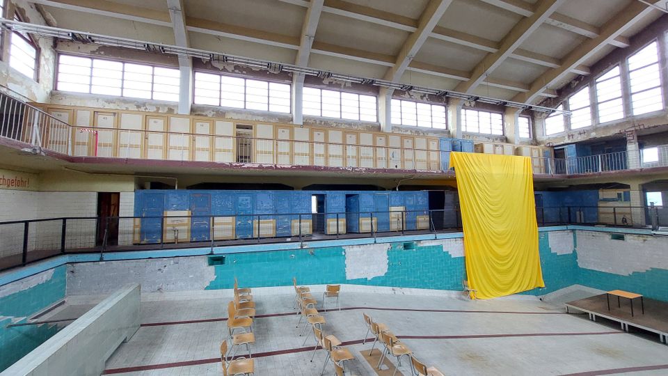 Ehemaliges Schwimmbad mit leerem Becken, abgeblätterter Wandfarbe und einer Galerie mit Umkleideschränken. Ein großes gelbes Tuch hängt von der Galerie, und im Beckenbereich stehen Stühle aufgestellt.