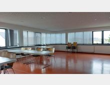 Ein heller, moderner Seminarraum mit orangefarbenem Boden, weißen Tischen und Stapelstühlen aus Holz. Große Fenster mit vertikalen Lamellenjalousien umgeben den Raum und lassen Tageslicht herein.