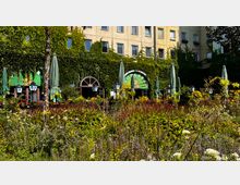 Außenbereich eines Restaurants mit grün bewachsener Fassade, großen Fenstern und geschlossenen Sonnenschirmen. Im Vordergrund befinden sich ein bunt bepflanztes Blumenbeet und Laternen, die den Eingangsbereich betonen.