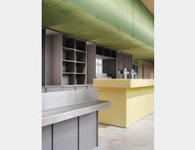 Minimalistisch gestalteter Innenraum mit einer grauen Regalwand, einer gelben Theke und einer Edelstahloberfläche. Die Decke ist mit einem grünen, durchscheinenden Paneel verkleidet, das Licht einfängt und reflektiert.