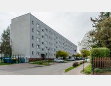 Mehrstöckiges Wohngebäude mit grauer Fassade und rechteckigen Fenstern, umgeben von einer Straßenkreuzung mit parkenden Autos und Gehwegen. Im Vordergrund befinden sich Bäume, ein Holzzaun und Straßenschilder.