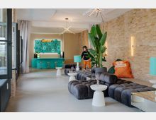 Moderner Lounge- und Barbereich mit freiliegender Ziegelwand, hohen Decken und türkisfarbener Theke samt Regalen im Hintergrund. Im Vordergrund stehen modulare, dunkelgraue Polstersofas mit kleinen weißen Beistelltischen, dazu große Zimmerpflanzen und filigrane Deckenleuchten.