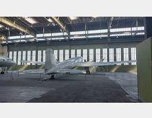 Großer Hangar mit einem silbernen Propellerflugzeug, das auf einem gepflasterten Boden präsentiert wird. Im Hintergrund befinden sich große grüne Tore mit vertikalen Fenstern, durch die Tageslicht einfällt.