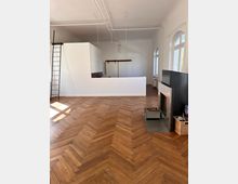 Wohnraum mit Fischgrätenparkett, einer modernen offenen Küche mit weißer Trennwand und einem Kamin mit Holzkorb im Vordergrund. Große Fensterbögen lassen Tageslicht ein und eine schwarze Leiter steht an der Wand.