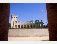 Blick auf eine historische Ruine mit erhaltenen Arkaden und einem quadratischen Turm aus Stein, eingerahmt von zwei roten Backsteinpfeilern im Vordergrund. Der blaue Himmel und umliegende Bäume bilden den Hintergrund der Szene.