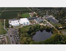 Luftaufnahme eines Industrie- oder Gewerbekomplexes mit mehreren weißen und grauen Gebäuden, großen Parkplätzen und einem kleinen See im Vordergrund, umgeben von Bäumen. Im Hintergrund befinden sich Felder und eine Wohngegend mit Gärten.