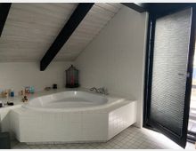 Badezimmer mit Eckbadewanne, gefliest mit weißen Kacheln, unter einer geneigten Holzbalkendecke. Neben der Wanne befindet sich ein dekorativer Vogelkäfig, und rechts ist eine Tür mit halb heruntergelassenem Plissee zu sehen.