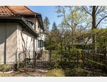 Seitenansicht eines Hauses mit hellem Putz und Ziegeldach, umgeben von einem Garten mit Bäumen und Sträuchern. Im Vordergrund ein schmiedeeisernes Tor mit einem bogenförmigen Eingang.