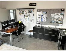 Modernes Büro mit minimalistischem Design. Ein Schreibtisch mit Computermonitoren und ergonomischem Stuhl, schwarze Schränke, eine Pinnwand sowie ein Projektionsbildschirm dominieren den Raum, ergänzt von grauem Fliesenboden und dezenten Deko-Elementen.