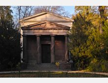 Klassizistisches Mausoleum mit säulenverzierter Fassade und dreieckigem Giebel, umgeben von hohen Bäumen in einer herbstlichen Parklandschaft. Die Eingangstür ist dunkel und mit Treppenstufen erreichbar.