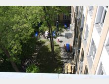 Innenhof eines Gebäudes mit gelblichen Backsteinfassaden und weißen Fenstern. Der Hof ist mit Bäumen, Sträuchern und Gehwegen gestaltet, enthält mehrere Tische und Bänke, teilweise mit blauen Tischdecken, sowie einige Personen, die zusammensitzen.