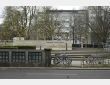 Städtischer Platz mit der Aufschrift "Theodor-Heuss-Platz" auf einer niedrigen Steinmauer, umgeben von Bäumen ohne Laub. Im Vordergrund stehen Fahrräder an einem Fahrradständer, während im Hintergrund moderne Gebäude zu sehen sind.