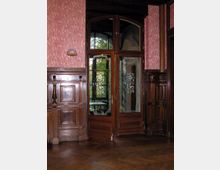 Holzvertäfelte Innenraumansicht mit einer doppelten Glastür, die mit dekorativen Mustern verziert ist. Der Raum hat rosafarbene Wände, einen Fischgrätparkettboden und sichtbare Spiegelungen von Bäumen im Glas der Tür.