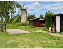 Eine Wiese mit einem kleinen Gebäude aus Holz, einer offenen Geräteschuppen-Tür rechts und einem runden, alten Aussichtsturm aus hellem Stein mit Metallleiter links im Hintergrund. Umgeben von Bäumen und Sträuchern, unter freiem Himmel mit leicht bewölktem Himmel.