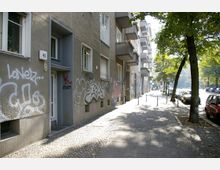 Straßenansicht eines Wohngebäudes mit grauer Fassade, auf der Graffiti aufgesprüht sind. Links der schmalen, mit Kopfsteinpflaster versehenen Gehweg stehen Bäume, während auf der rechten Straßenseite parkende Autos und eine belebte Straße zu sehen sind.