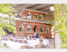 Holzgebäude mit der Aufschrift „Oberhafen-Kantine“, das unter einer Betonüberdachung steht. Vor dem Gebäude sind mehrere gedeckte Tische mit weißen Tischdecken und Blumen sowie dekorative Luftballons und hängende Papierdekorationen zu sehen.