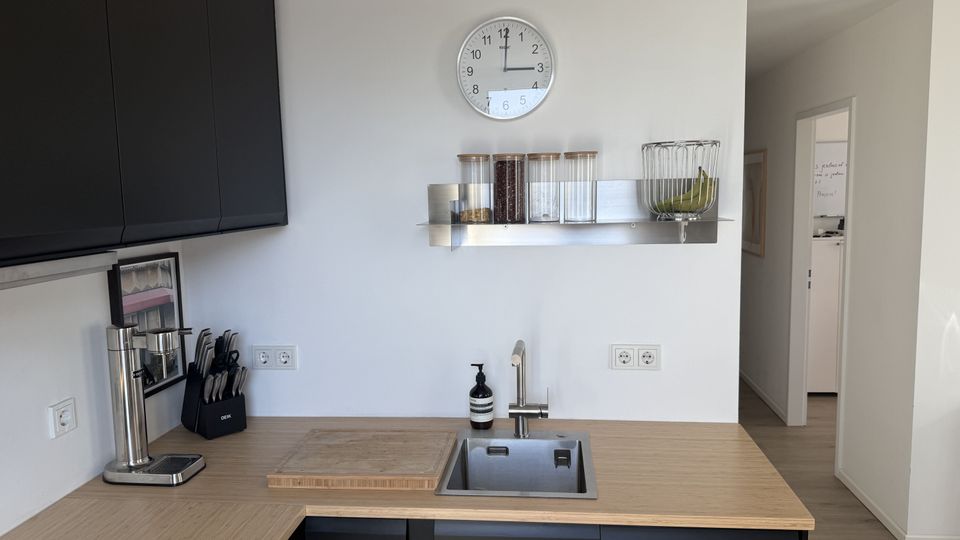 Moderne Küchenzeile mit Holzarbeitsplatte und eingelassenem Edelstahlspülbecken, darüber eine Wanduhr und ein Metallregal mit Vorratsgläsern und Obstkorb. Links sind dunkle Hängeschränke, eine Kaffeemaschine und ein Messerblock zu sehen; rechts führt ein Durchgang in einen hellen Flur.