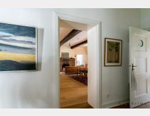 Innenansicht eines Hauses mit Blick durch eine offene Tür in ein Wohnzimmer. Im Vordergrund weiße Wände mit zwei Gemälden und einer halb geöffneten Tür; im Hintergrund ein Raum mit Holzboden, einem Esstisch und Stühlen sowie einer Couch vor einem gemauerten Kamin.