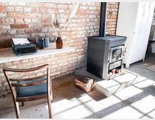 Ein rustikaler Innenraum mit einer freigelegten Ziegelwand, einem Holzofen mit gestapeltem Brennholz daneben, einem schlichten Holztisch mit einer Schreibmaschine und Büchern darauf sowie einem Holzstuhl. Im Hintergrund steht ein weißer Metallschrank.