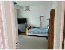 Blick in ein Kinderzimmer mit weiß gestrichenen Wänden, einem Einzelbett mit blau gemustertem Bettzeug, einer grauen Kommode mit Fernseher darauf und einem Hängelampenschirm. Der Boden besteht aus Holzdielen, teils bedeckt mit einem hellen Teppich.