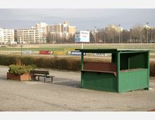 Grün gestrichener Kiosk und Sitzbank neben einem gemauerten Pflanzbehälter auf einem gepflasterten Bereich vor einer Pferderennbahn; im Hintergrund Wohngebäude und Stehtribünen.
