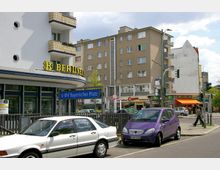 Straßenszene am Bayerischen Platz in Berlin mit modernen Wohngebäuden, Geschäften, einer Bankfiliale und der U-Bahn-Eingangsbeschilderung "U-Bhf Bayerischer Platz". Im Vordergrund sind parkende Autos und eine Kreuzung mit Ampeln zu sehen.