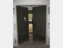 Geöffnete schwere Doppeltür aus dunklem Metall in einem einfachen, weißen Gebäudeeingang; dahinter ein Außenbereich mit einem geparkten blauen Auto und Bäumen im Hintergrund.