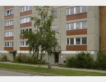 Mehrgeschossiges Wohngebäude aus Betonplatten mit roten Zierelementen, mehreren weißen Fenstern und einem zentralen Eingang. Davor befindet sich ein kleiner Gehweg, ein schmaler Grünstreifen sowie ein einzelner Baum.