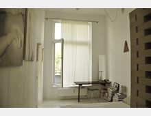Helles Schlafzimmer mit weiß gestrichenen Wänden, einem großen Fenster mit halb geöffnetem Vorhang und minimalistischen Möbeln. Ein Schreibtisch mit einem Stuhl, ein Stapel Bücher und ein Standspiegel befinden sich an der Wand, daneben hängen Kunstwerke.