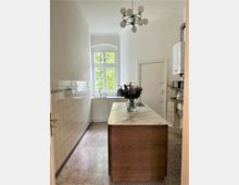 Schmale, helle Küche mit hohen Decken, einer Holzinsel mit Marmorarbeitsplatte und einer Vase mit Blumen. Im Hintergrund ein großes Fenster mit Blick ins Grüne, helle Fliesen an den Wänden und ein moderner Kronleuchter an der Decke.