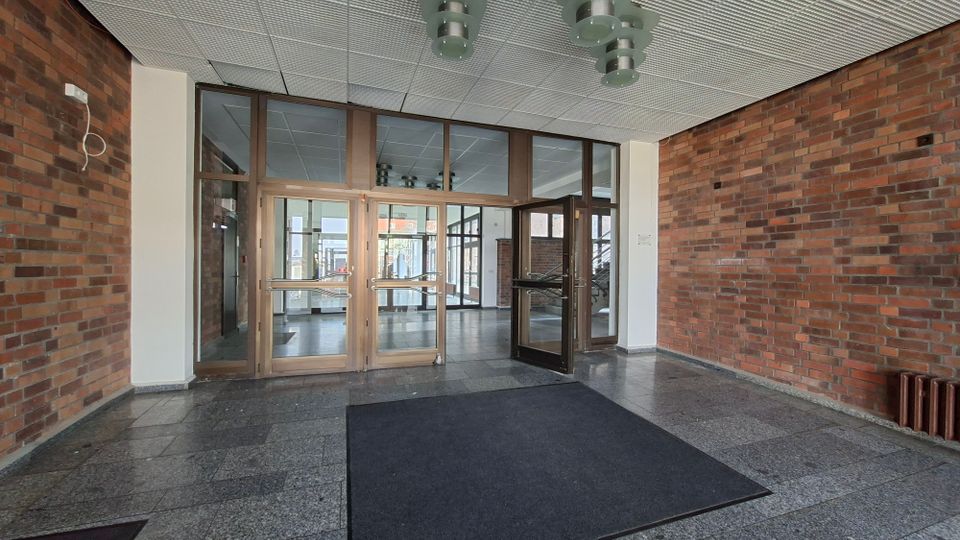Eingangsbereich eines Gebäudes mit Backsteinwänden, grauem Steinboden und Rasterdecke mit Deckenleuchten. Im Zentrum stehen große verglaste Türen und Fensterrahmen, die in eine helle Lobby mit weiteren Glasfronten führen.